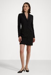 Žena stojí oblečená v čiernom sakošate s gombíkmi a dlhými rukávmi, v čiernych sandáloch na opätku, pred jednoduchým pozadím.