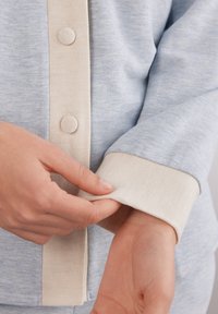 Hellblaue Bluse aus weichem Stoff mit einer cremefarbenen Knopfleiste und Manschetten. Die Manschettentextur ist glatt, mit runden Stoffknöpfen.