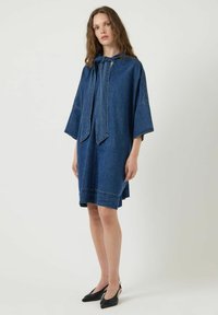 Vestido de mezclilla en azul oscuro, con un corte holgado, mangas anchas y un lazo en el escote. Textura suave con detalles de costura visibles.