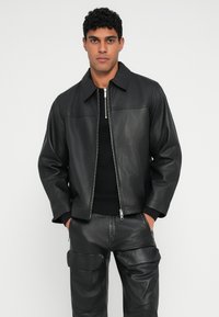 Chaqueta de cuero negra con un collarín puntiagudo, cierre de cremallera y dos bolsillos frontales, combinada con pantalones de cuero negro a juego. Textura suave.