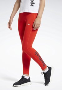 Czerwone sportowe legginsy o dopasowanym kroju, z czarnym logo "Reebok" i gładką, elastyczną fakturą. W zestawie z czarnymi sneakersami.