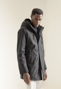 Parka impermeable negra con capucha, con bolsillos delanteros, cremallera y cierres de presión. La tela tiene una textura suave.