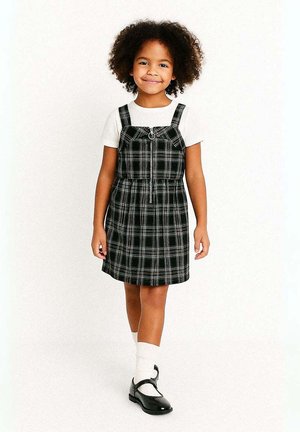 Fille aux cheveux bouclés marchant, portant un t-shirt blanc, une robe à carreaux noire et blanche avec fermeture éclair, des chaussettes blanches et des chaussures Mary Jane noires.