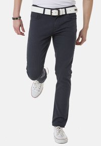 Marineblaue, strukturierte Chinos mit schlankem Schnitt, ausgestattet mit einem weißen Gürtel, kombiniert mit weißen Sneakers mit roten Akzenten.