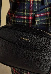Sac Joules en cuir texturé noir avec fermeture éclair porté sur un vêtement en tissu tartan à carreaux verts et rouges.