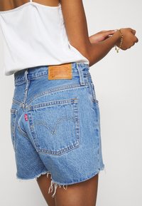 Donna che indossa pantaloni corti di jeans azzurri con orli sfrangiati e un top bianco senza maniche, mentre aggiusta un bracciale di catena dorato al polso destro.