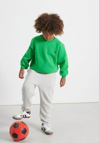 Felpa verde con polsini a costine, pantaloni grigi chiari e sneakers bianco e nere, mentre dribbla un pallone da calcio nero e rosso.