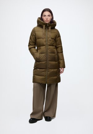 Femme portant un long manteau matelassé à capuche vert olive avec fermeture éclair devant, pantalon marron à jambes larges et chaussures noires, debout sur un fond blanc.