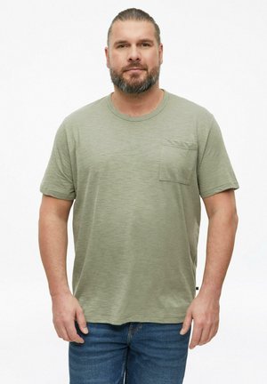 Man met baard en naar achter gebonden haar, gekleed in een lichtgroen T-shirt met korte mouwen en borstzak en een blauwe spijkerbroek, staand tegen een witte achtergrond.