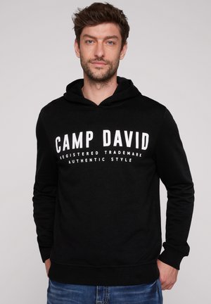 Mężczyzna w czarnej bluzie z kapturem z napisem "CAMP DAVID" i niebieskich dżinsach stoi na prostym jasnym tle.