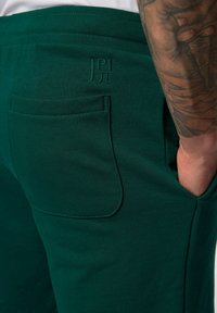 Zelené bavlněné joggery mají boční kapsu a embosovaný design loga. Tkanina vypadá měkce a má hladkou texturu.