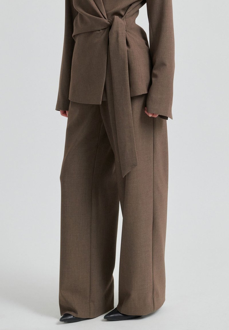 Pantalon marron à jambes larges associé à un blazer assorti ceinturé et des chaussures noires à bout pointu sur un fond clair uni.