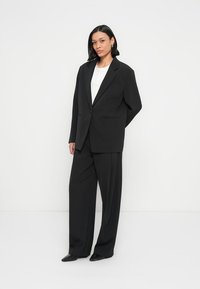 Costume noir sur mesure avec une veste à un bouton et un pantalon large, présentant une texture lisse et un design minimaliste. Haut blanc en dessous.