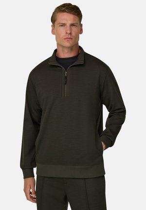 Man draagt een donkergroene pullover met kwartrits en bijpassende broek, staand met één hand in de zak tegen een effen achtergrond.