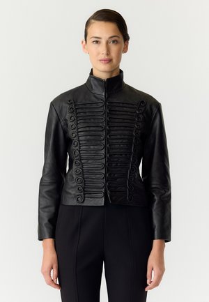 Une femme présente une veste en cuir noire à col montant et ornée de boucles tressées décoratives, portée avec un pantalon noir, sur un fond uni.