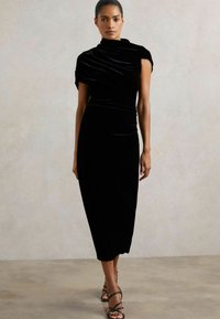 Čierne zamatové šaty s priliehavým strihom, asymetrickým dizajnom ramena a nariasenými detailmi, v kombinácii s čiernymi sandálmi na opätku.