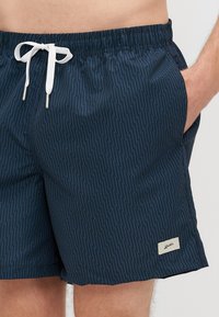 Tumesinised ujumispüksid kalavõrkpaariga mustriga, elastse vöökoha, valge paela ja küljetaskutega. Omab väikest brändilogo ääres.