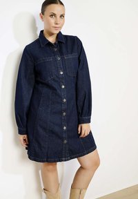Donkerblauwe denim jurk met lange mouwen, knoopsluiting aan de voorkant en twee borstzakken. Heeft een ontspannen fit en lichte stikdetails.