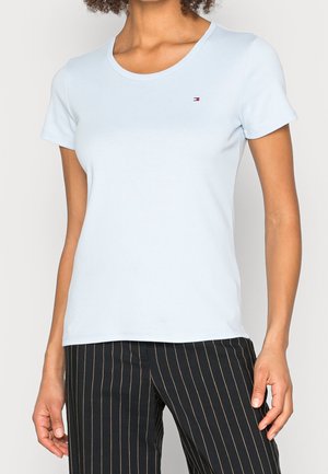 Mujer con una camiseta de manga corta azul claro con un pequeño logo en el pecho y pantalones negros con rayas finas.