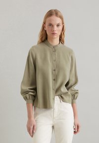 Olijfgroene blouse met knopen, een staande kraag, opgezette mouwen en een ontspannen pasvorm. Gemaakt van een gladde stof met een subtiele glans.