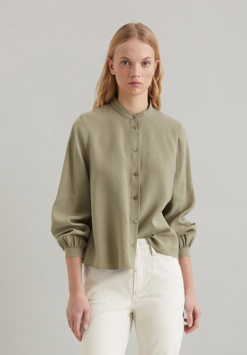 Olijfgroene blouse met knopen, een staande kraag, opgezette mouwen en een ontspannen pasvorm. Gemaakt van een gladde stof met een subtiele glans.