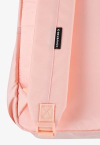 Sac à dos en tissu rose avec une texture lisse, arborant une étiquette noire Converse, des bretelles réglables et un détail de fermeture à boucle.