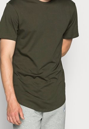 Mand iført en almindelig mørkegrøn kortærmet t-shirt og lysegrå joggingbukser, står med den ene hånd bag ryggen.