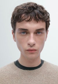 Ritratto di una persona con capelli corti e ondulati di colore castano, occhi azzurri, che indossa un maglione testurizzato beige chiaro sopra una camicia nera. Sfondo neutro.