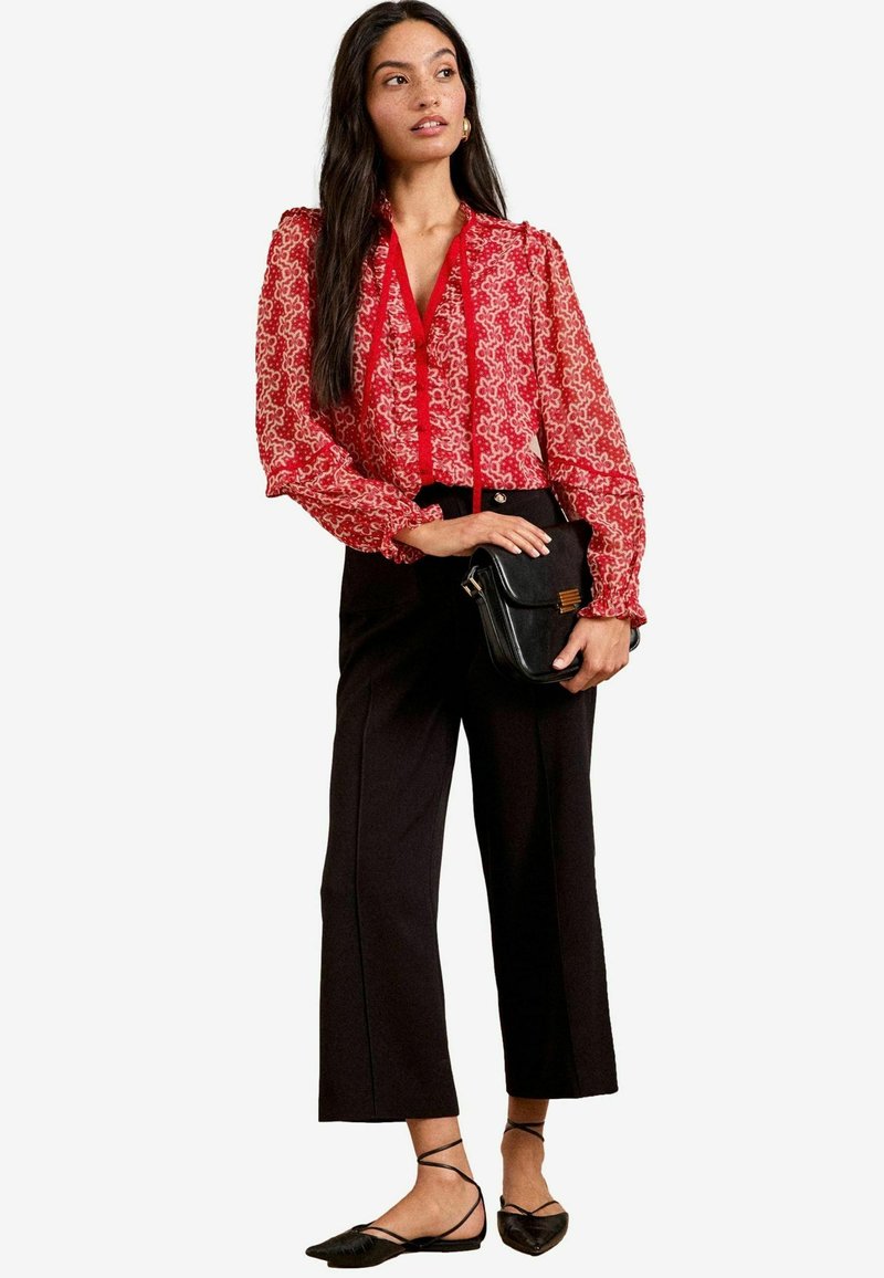 Blusa rossa a fantasia con maniche lunghe e voluminose, abbinata a pantaloni neri a vita alta e gamba larga e scarpe basse nere con cinturino.
