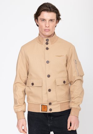 Mężczyzna w beżowej, zapinanej na guziki kurtce bomberce z naszywką i kieszenią na zamek, stojący na białym tle.