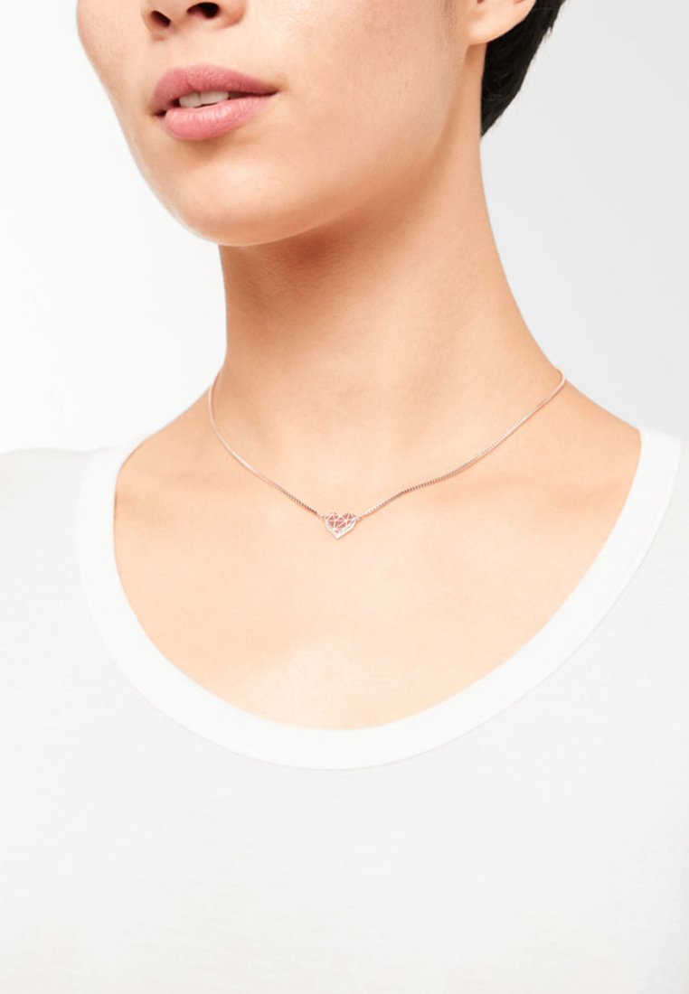 Zilveren hartketting met een geometrisch ontwerp, hangend aan een delicate ketting, gedragen boven een eenvoudig wit shirt.