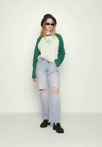 T-shirt raglan verde e creme com gráfico "RAGGED", jeans azul desgastado e sapatos de plataforma pretos. Modelo usa um chapéu preto e óculos de sol.