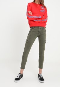 Röd sweatshirt med grafisk text på ärmarna, olivgröna cargobyxor med flera fickor och svarta sneakers med vita detaljer.