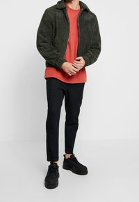 Groene jas met een pluizige textuur en ritssluiting, gecombineerd met een rood T-shirt en zwarte slim-fit broek. Zwarte schoenen met een dikke zool.