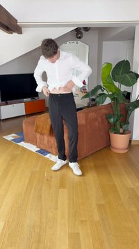 Hvit langermet skjorte, sorte bukser med knapp og snøring, person som justerer midjebåndet, innendørs setting med planter og møbler.