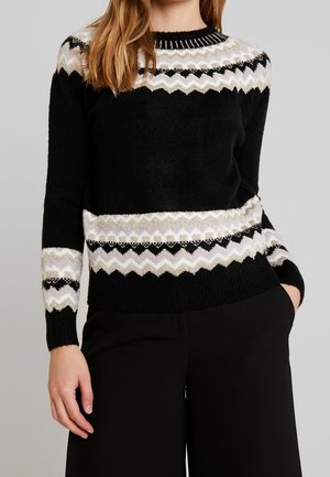 Kobieta w czarnym dzianinowym swetrze z białymi, beżowymi i czarnymi wzorami zygzakowymi na klatce piersiowej, rękawach i talii, w połączeniu z czarnymi spodniami.