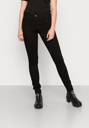 Persona usando jeans ajustados negros y botas de tobillo negras con detalle de cremallera plateada, de pie frente a un fondo blanco.