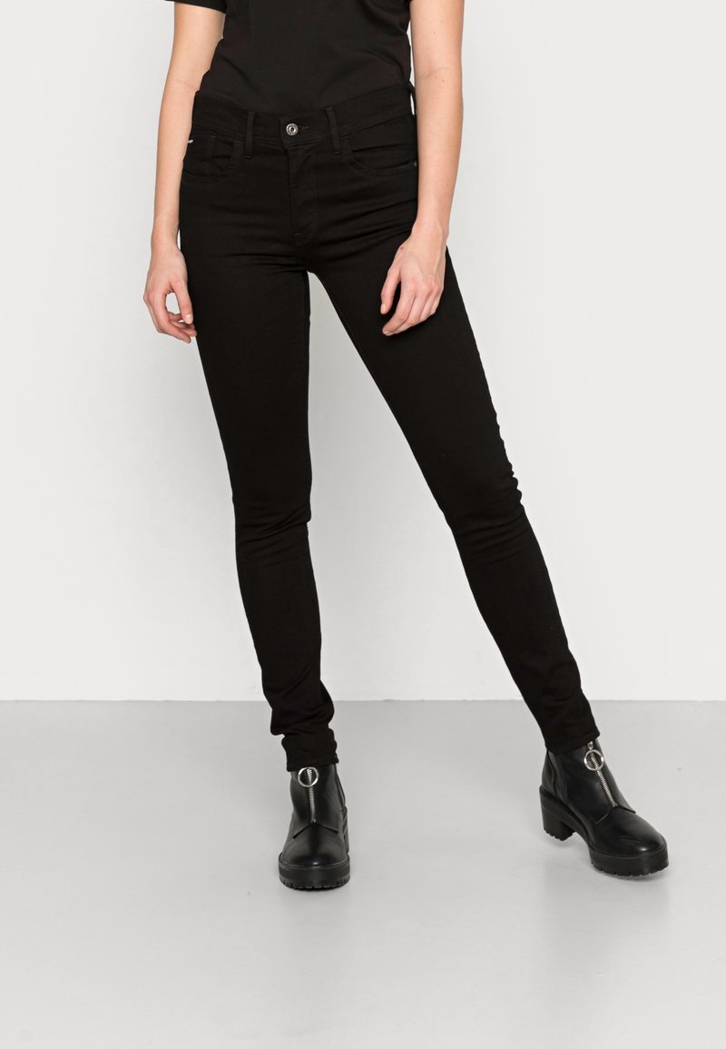 Persona usando jeans ajustados negros y botas de tobillo negras con detalle de cremallera plateada, de pie frente a un fondo blanco.