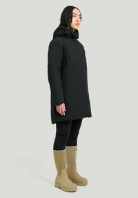 Svart vattentät parka med huva, med rak passform och långa ärmar, tillsammans med beiga gummistövlar.