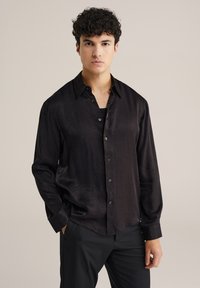 Noor mehine juustega noormees, kes kannab läikivat musta nööpidega särk ja musti pükse, seistes ühe käega taskus lihtsa tausta ees.