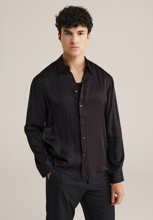 Mladý muž s kučeravými vlasmi, oblečený v lesklých čiernych košeliach s gombíkmi a čiernych nohaviciach, stojí s jednou rukou v vrecku na jednotnom pozadí.