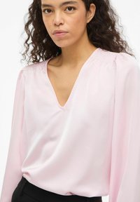 Blouse en satin rose avec un col en V et des manches longues, présentant des détails froncés aux épaules et une texture lisse.