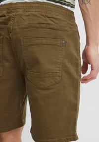 Pantaloni scurți din bumbac verde măsliniu, cu croială lejeră. Au buzunare la spate cu un model discret de cusături și detalii metalice rivetate.