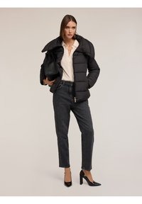 Giacca imbottita nera con un grande colletto, abbinata a una blouse chiara e jeans scuri con una striscia laterale. Scarpe nere con tacco alto e borsa.