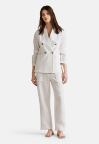 Blazer de lino blanco de doble botonadura con botones marrones, combinado con pantalones de lino de corte relajado que tienen bolsillos de parche. Textura suave.