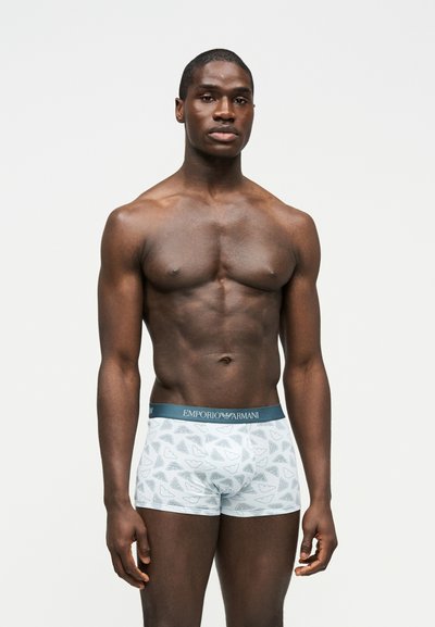 Muskularny mężczyzna stojący, ubrany w białe i szare bokserki Emporio Armani z wzorem, na jednolitym jasnym tle.