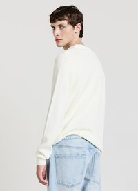 Maglione bianco lavorato a maglia con motivo a rilievo, vestibilità morbida e polsini a coste, abbinato a jeans denim azzurri. La modella è leggermente girata.