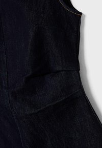 Tumman denim-kangas, jossa on sileä tekstuuri. Sisältää kaarevia viivoja ja kontrastista oranssia ommelta. Mukana on pieni taskudetaili sivulla.