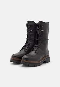 Botas de combate de cuero negro con suelas gruesas y estriadas, cordones frontales, ojales de metal, cremallera lateral y ribete marrón alrededor de la suela.