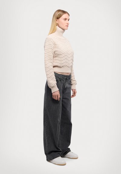 Lichtbeige, textuurgebreide coltrui met een zigzagpatroon, gecombineerd met loszittende, high-waisted donkergrijze jeans en witte sneakers.